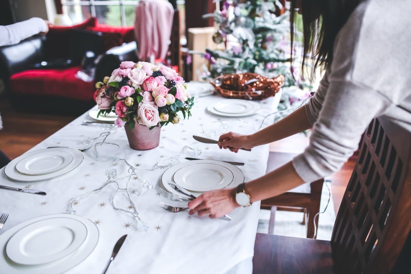 A woman sets a dinner table at home with white and gold place settings and pink flowers